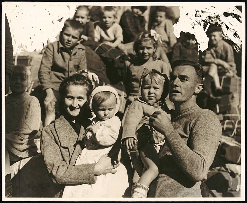 Eine große Gruppe an Menschen ist zu sehen, links im Vordrergrund eine lachende Frau, zwei Kinder in der Mitte und rechts ein lachender Mann.