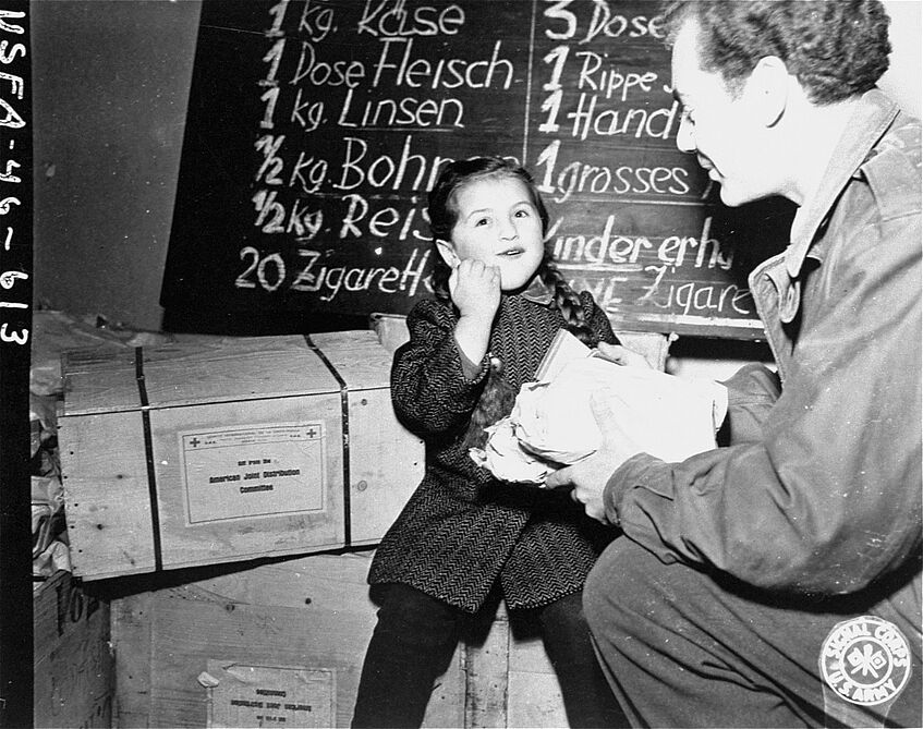 Ein junges Mädchen sitzt auf Kisten (des roten Kreuzes?). Ein Soldat gibt ihr Essen. Das Kind lacht.
